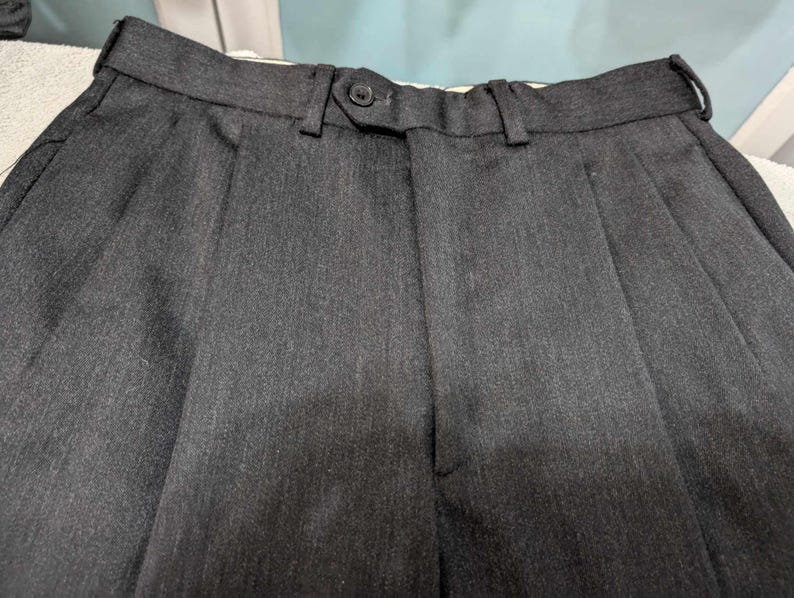 Puede incluir: Primer plano de un pantal&oacute;n plisado gris oscuro. El pantal&oacute;n tiene cierre de bot&oacute;n y trabillas para el cintur&oacute;n. La tela tiene una sutil textura vertical. Los pantalones probablemente forman parte de un traje o vestimenta formal.