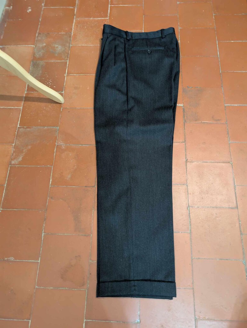 Puede incluir: Pantalones de vestir gris oscuro con dobladillo vuelto. Los pantalones tienen cierre de bot&oacute;n y trabillas para el cintur&oacute;n. La tela tiene un sutil patr&oacute;n de rayas verticales. Los pantalones est&aacute;n doblados cuidadosamente sobre una superficie embaldosada.