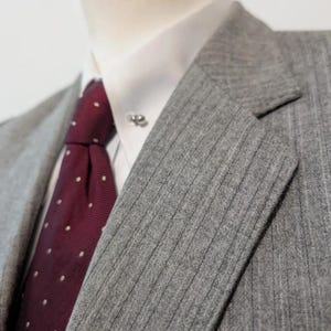 May include: Close-up of a gray pinstripe suit jacket with a white dress shirt and a burgundy tie with white polka dots. A silver tie pin is visible on the collar. The suit jacket has a notched lapel.