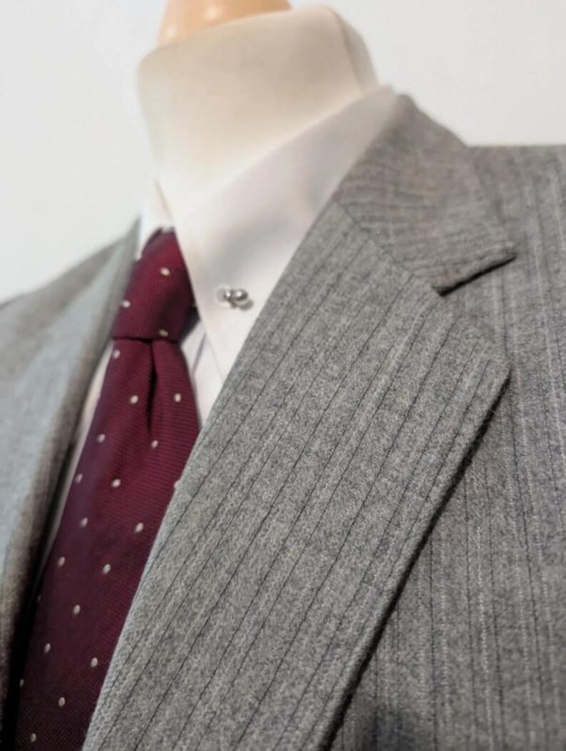 May include: Close-up of a gray pinstripe suit jacket with a white dress shirt and a burgundy tie with white polka dots. A silver tie pin is visible on the collar. The suit jacket has a notched lapel.