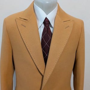Puede incluir: Un abrigo de color beige de doble botonadura con solapa de muesca. El abrigo se combina con una camisa blanca y una corbata estampada en burdeos y blanco. El abrigo est&aacute; confeccionado con costuras visibles.