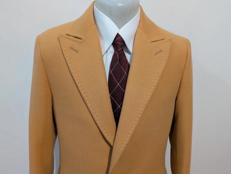 Puede incluir: Un abrigo de color beige de doble botonadura con solapa de muesca. El abrigo se combina con una camisa blanca y una corbata estampada en burdeos y blanco. El abrigo est&aacute; confeccionado con costuras visibles.