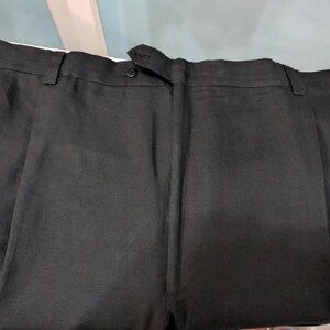 May include: Close-up of black dress pants with a button closure and pleats. The pants are made of a dark fabric and appear to be tailored. The image shows the waistband and the top portion of the pants.