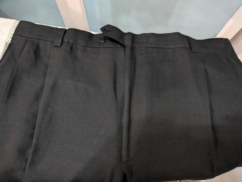 May include: Close-up of black dress pants with a button closure and pleats. The pants are made of a dark fabric and appear to be tailored. The image shows the waistband and the top portion of the pants.