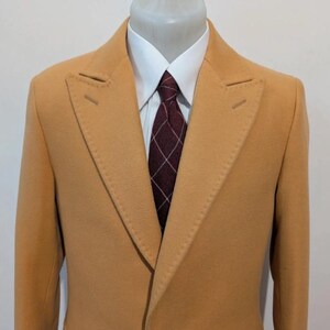 Puede incluir: Un abrigo color beige con solapa de muesca, camisa blanca y corbata estampada en burdeos y blanco. El abrigo se exhibe en un maniqu&iacute;. El abrigo tiene dos aberturas decorativas en la solapa.