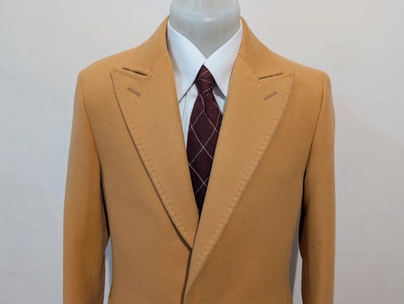 Puede incluir: Un abrigo color beige con solapa de muesca, camisa blanca y corbata estampada en burdeos y blanco. El abrigo se exhibe en un maniqu&iacute;. El abrigo tiene dos aberturas decorativas en la solapa.