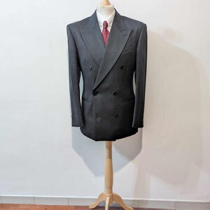 Puede incluir: Chaqueta de traje cruzada gris antracita, camisa blanca con cuello y corbata roja estampada. La chaqueta tiene dos filas de botones y se exhibe en un maniqu&iacute; de madera.