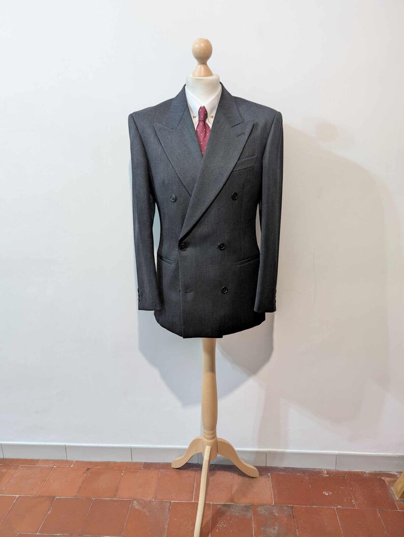 Puede incluir: Chaqueta de traje cruzada gris antracita, camisa blanca con cuello y corbata roja estampada. La chaqueta tiene dos filas de botones y se exhibe en un maniqu&iacute; de madera.