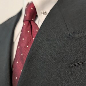 May include: Close-up of a black suit jacket with a white dress shirt and a burgundy tie. The tie features a white polka dot pattern. A silver tie pin is visible on the shirt collar. The suit jacket has a notched lapel and a single button.