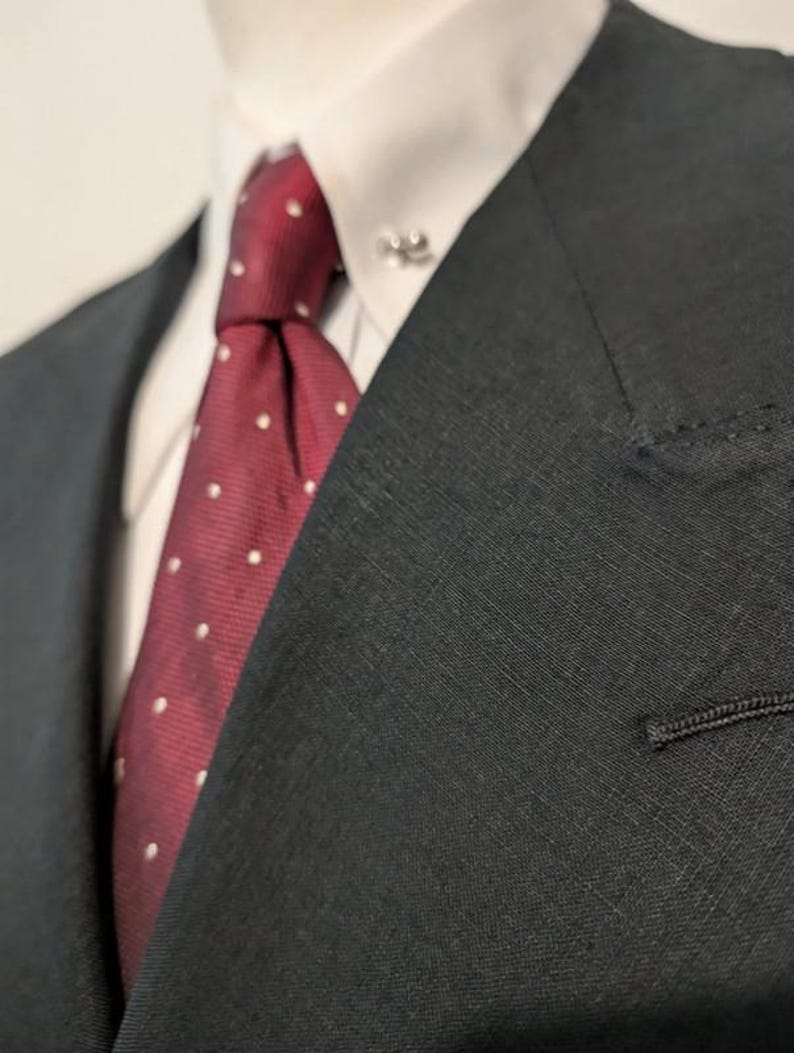 May include: Close-up of a black suit jacket with a white dress shirt and a burgundy tie. The tie features a white polka dot pattern. A silver tie pin is visible on the shirt collar. The suit jacket has a notched lapel and a single button.