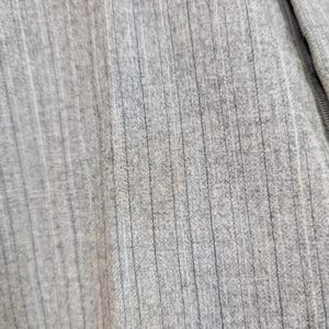 May include: Close-up of a light gray ribbed knit fabric. The fabric has vertical lines and a soft texture. The material appears to be a blend of fibers, possibly cotton or a similar material. The image shows a section of a garment.