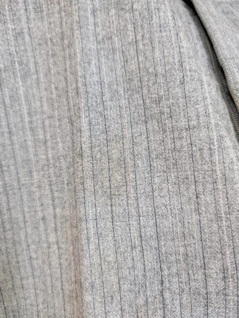 May include: Close-up of a light gray ribbed knit fabric. The fabric has vertical lines and a soft texture. The material appears to be a blend of fibers, possibly cotton or a similar material. The image shows a section of a garment.