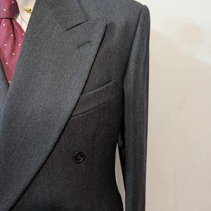 Puede incluir: Chaqueta de traje cruzada gris carb&oacute;n con solapas con muescas. La chaqueta tiene dos filas de botones y un bolsillo ribeteado. Se ven una camisa blanca y una corbata burdeos con lunares blancos.