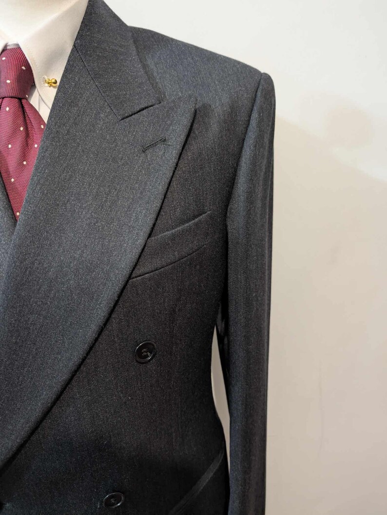 Puede incluir: Chaqueta de traje cruzada gris carb&oacute;n con solapas con muescas. La chaqueta tiene dos filas de botones y un bolsillo ribeteado. Se ven una camisa blanca y una corbata burdeos con lunares blancos.