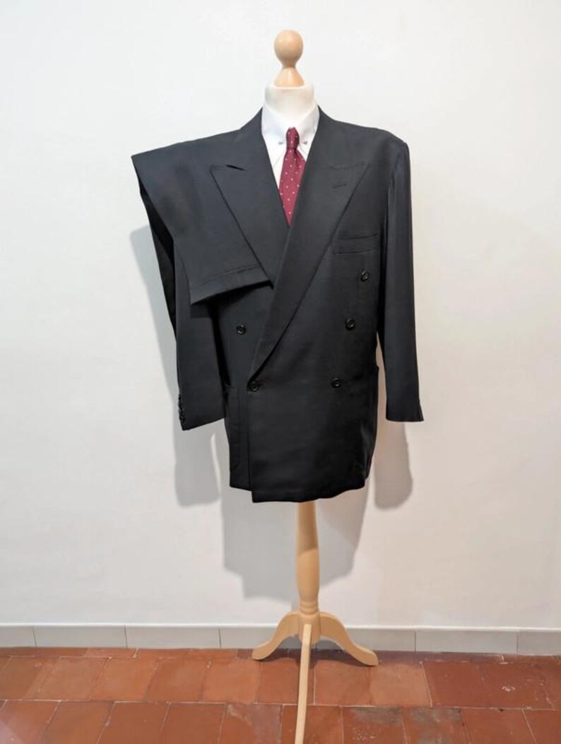 May include: A black double-breasted suit displayed on a wooden mannequin. The suit includes a white collared shirt and a red patterned tie. The mannequin is on a wooden stand, set against a white wall and a terracotta tile floor.