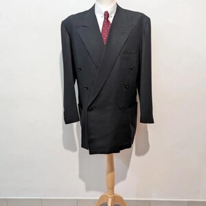 May include: A black double-breasted blazer with a white collared shirt and a red patterned tie. The blazer is displayed on a wooden mannequin. The background is a plain white wall and a terracotta tile floor.