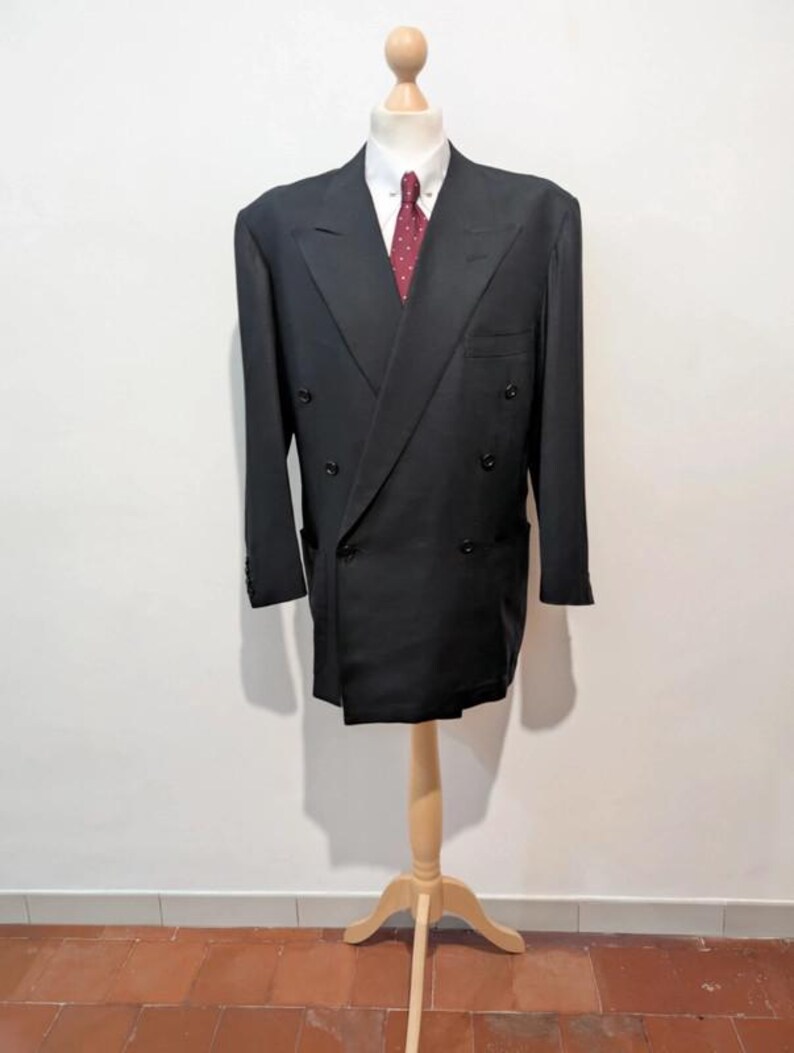 May include: A black double-breasted blazer with a white collared shirt and a red patterned tie. The blazer is displayed on a wooden mannequin. The background is a plain white wall and a terracotta tile floor.