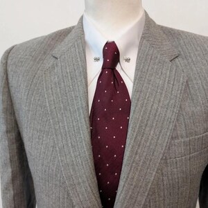 May include: A gray pinstripe suit jacket with a burgundy tie featuring white polka dots. The suit is worn over a white dress shirt with decorative collar pins. The jacket has a notched lapel and a pocket.