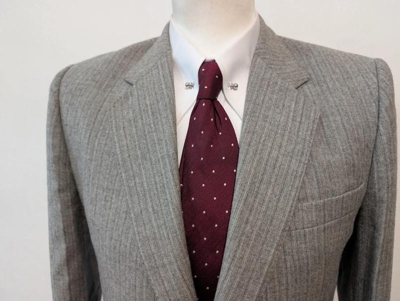 May include: A gray pinstripe suit jacket with a burgundy tie featuring white polka dots. The suit is worn over a white dress shirt with decorative collar pins. The jacket has a notched lapel and a pocket.
