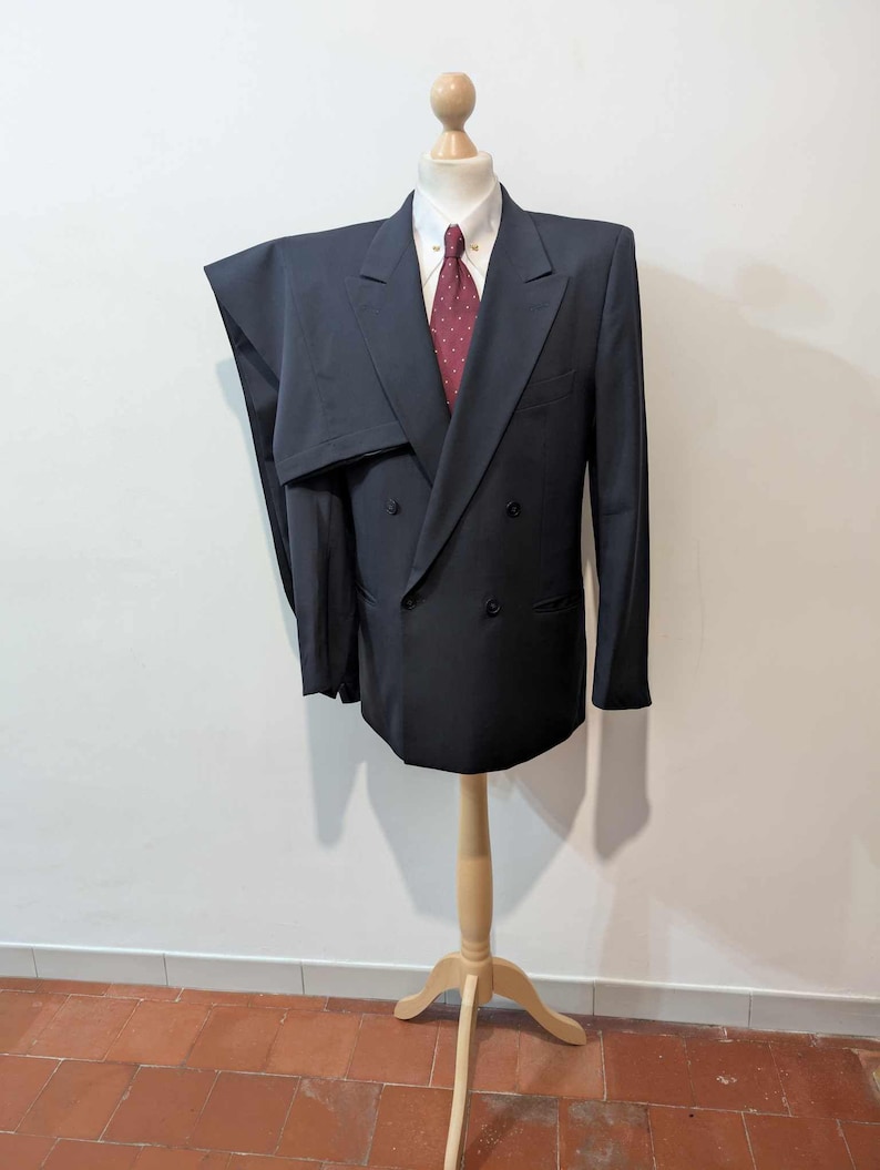May include: A navy blue double-breasted suit on a wooden mannequin. The suit comprises a jacket and trousers, worn with a white shirt and a burgundy tie with white spots. The mannequin is on a wooden stand. The suit is a classic formal wear.