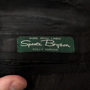 May include: Close-up of a black garment with a green label. The label reads "Pure Irish Linen" and "Spence Bryson" in white script. The words "Fully Shrunk" are also visible on the label.