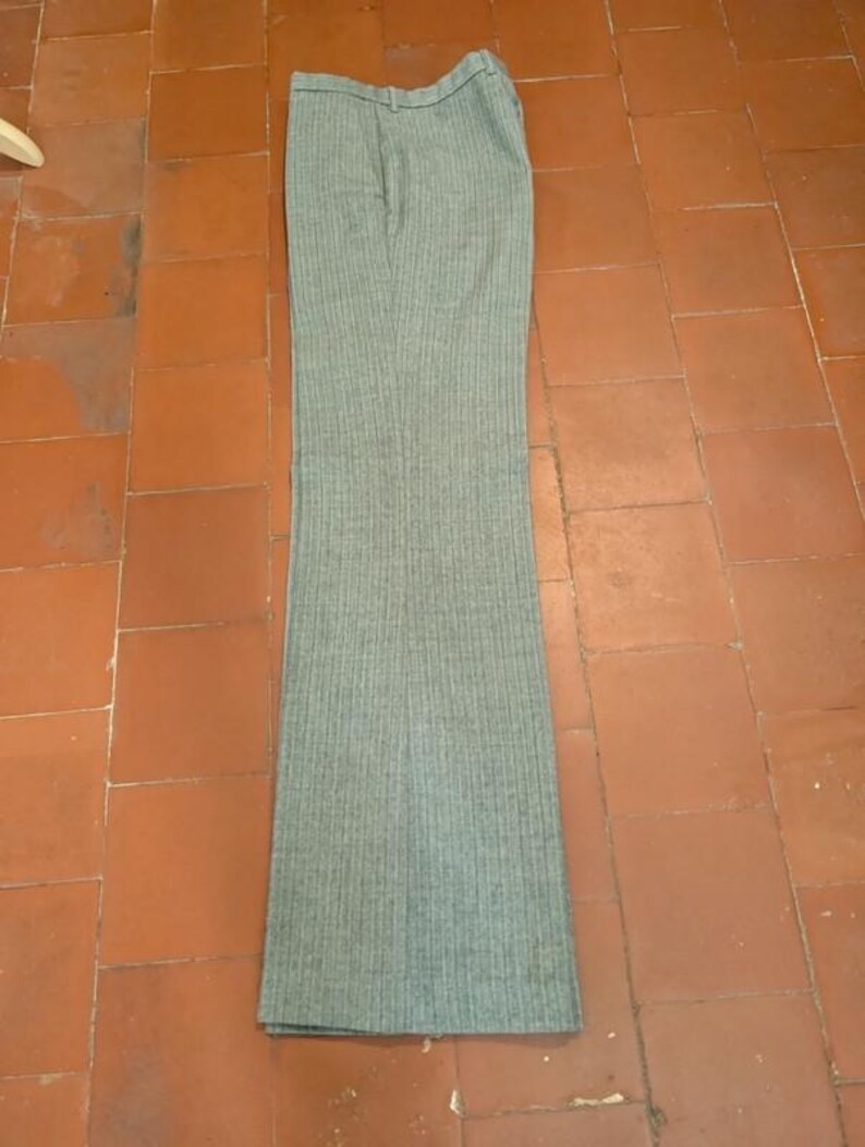 May include: A pair of gray, vertically striped trousers. The pants have a high waist and a flared leg. The fabric appears to be a medium-weight material, suitable for cooler weather. The trousers are laid flat on a tiled surface.