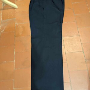 May include: Navy blue tailored trousers. The trousers are made from a smooth fabric and have a classic design. They feature a button and zip fastening, pockets, and a cuffed hem. The trousers are laid flat on a tiled surface.