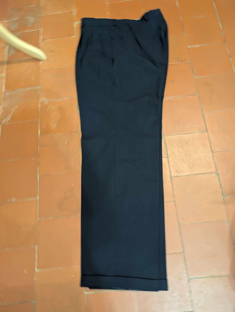May include: Navy blue tailored trousers. The trousers are made from a smooth fabric and have a classic design. They feature a button and zip fastening, pockets, and a cuffed hem. The trousers are laid flat on a tiled surface.