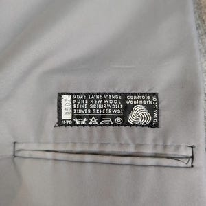 May include: Close-up of a gray fabric with a stitched pocket. A black and white label is sewn onto the fabric, displaying text including "PURE NEW WOOL" and a Woolmark logo. The fabric appears to be part of a garment.