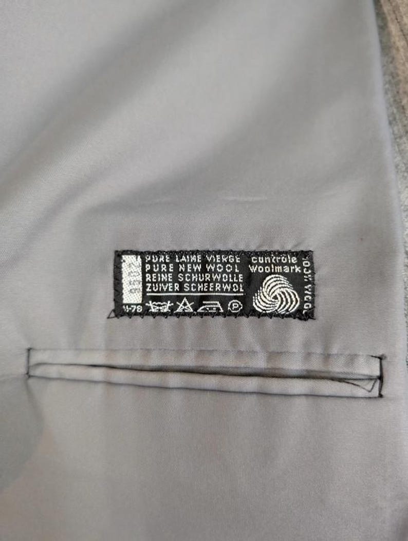 May include: Close-up of a gray fabric with a stitched pocket. A black and white label is sewn onto the fabric, displaying text including "PURE NEW WOOL" and a Woolmark logo. The fabric appears to be part of a garment.