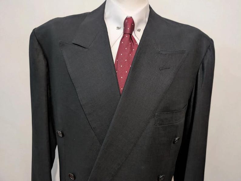 May include: A black double-breasted suit jacket with a white dress shirt and a burgundy tie with white polka dots. The jacket has a notched lapel and a pocket on the left side. The suit is displayed on a mannequin.