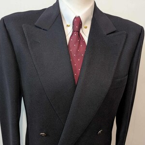 May include: A dark navy double-breasted blazer with a notched lapel, worn with a white dress shirt and a burgundy tie with white polka dots. The blazer has two rows of buttons and a pocket on the left side.