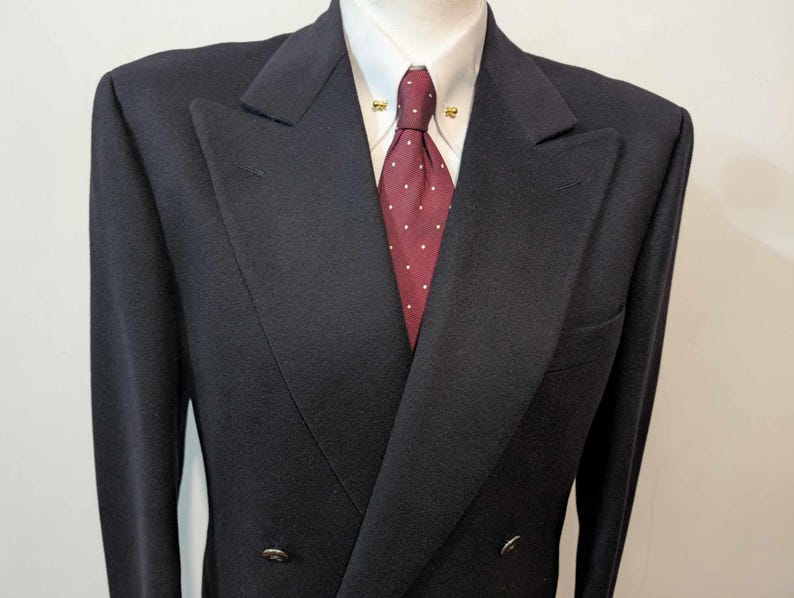 May include: A dark navy double-breasted blazer with a notched lapel, worn with a white dress shirt and a burgundy tie with white polka dots. The blazer has two rows of buttons and a pocket on the left side.