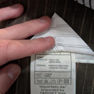 May include: Close-up of a brown pinstripe fabric with a white lining. A tag is visible with text in Italian and English, including fabric composition and care instructions. The tag also includes the importer's information.