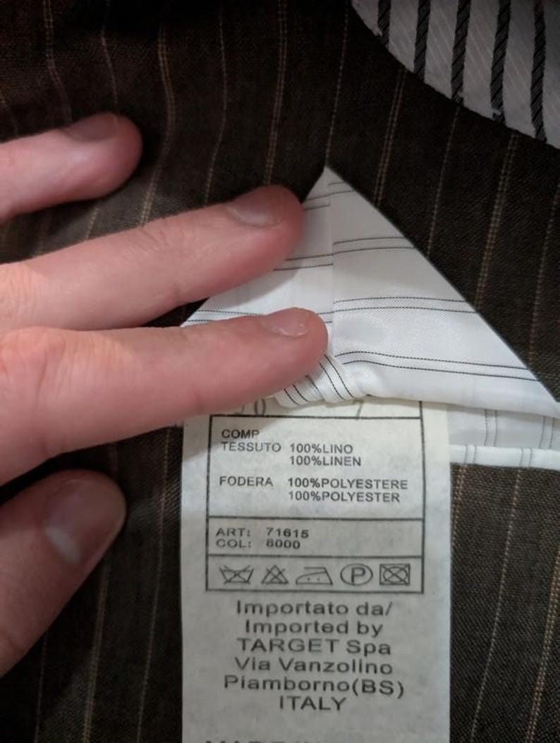 May include: Close-up of a brown pinstripe fabric with a white lining. A tag is visible with text in Italian and English, including fabric composition and care instructions. The tag also includes the importer's information.