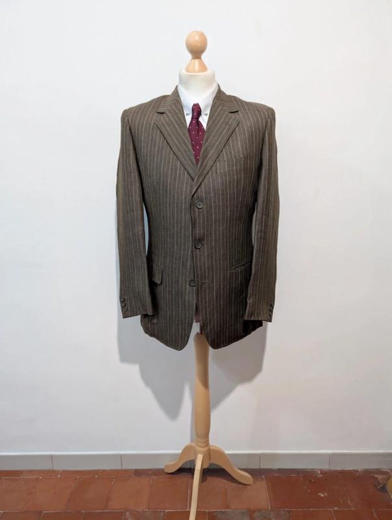 May include: A brown pinstripe blazer with a white dress shirt and a burgundy polka dot tie. The three-button blazer is displayed on a wooden mannequin. The blazer has two front pockets and three buttons on each sleeve.