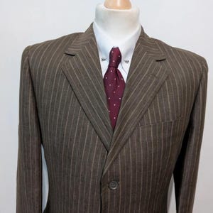 May include: A brown pinstripe suit jacket with a white collared shirt and a burgundy tie with white polka dots. The jacket has a single button closure and a chest pocket. The suit is displayed on a mannequin.