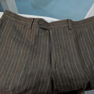 May include: Brown pinstripe trousers with a button closure and belt loops. The trousers are made of a textured fabric and have vertical stripes in a lighter shade of brown. The pants are folded.