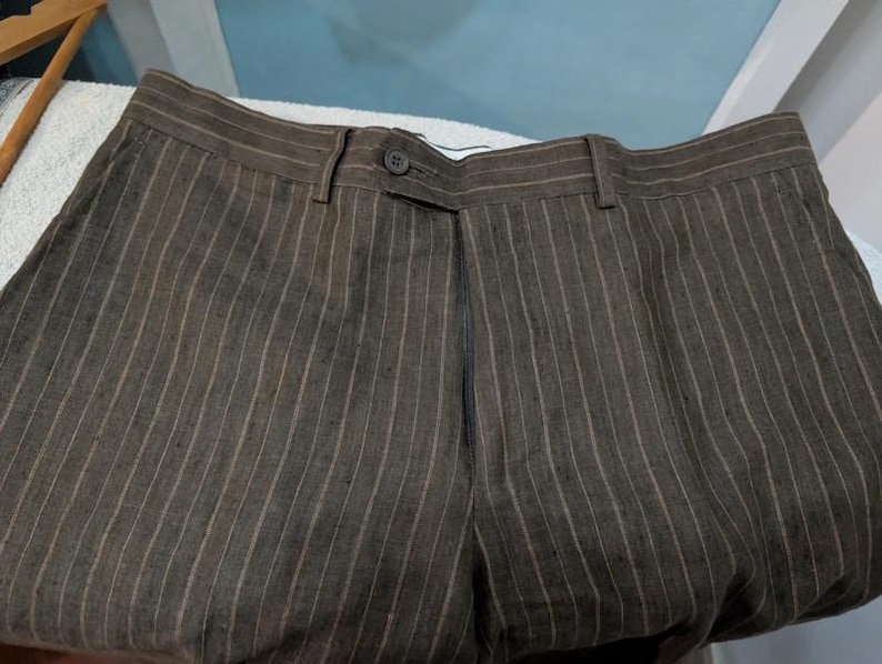 May include: Brown pinstripe trousers with a button closure and belt loops. The trousers are made of a textured fabric and have vertical stripes in a lighter shade of brown. The pants are folded.