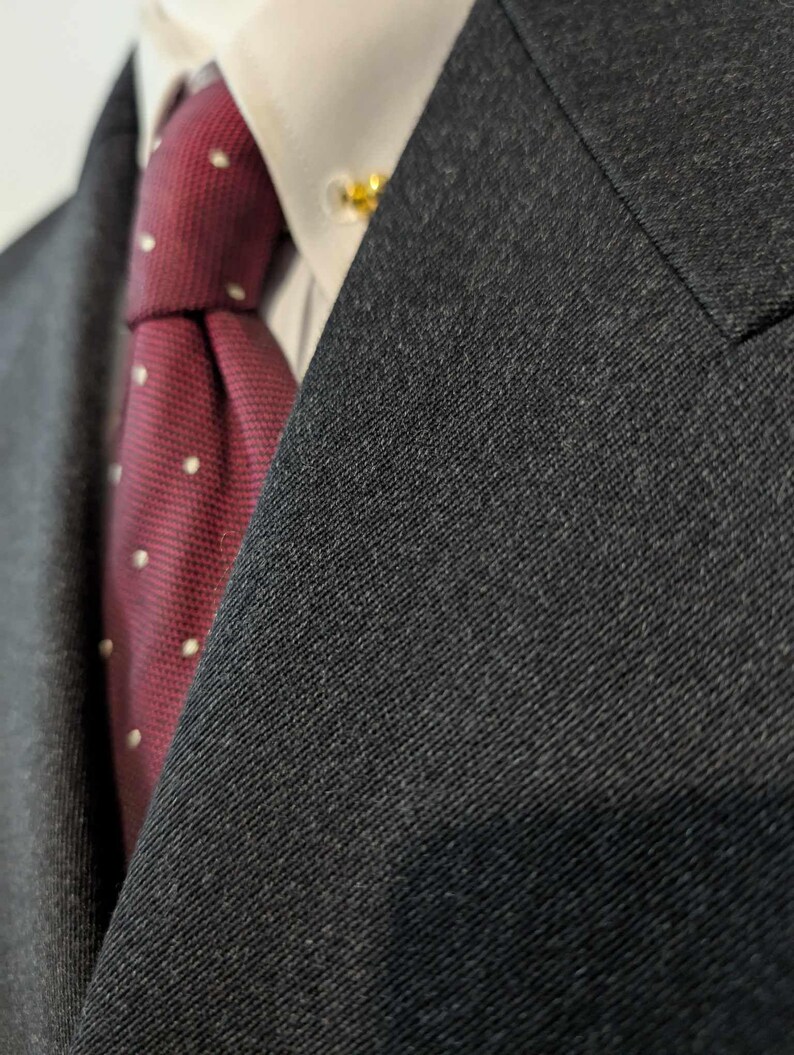 Puede incluir: Primer plano de una chaqueta de traje gris oscuro con una camisa blanca con cuello y una corbata burdeos. La corbata presenta peque&ntilde;os lunares blancos. Se ve un alfiler de corbata dorado.