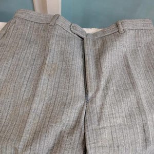 May include: Close-up of gray pinstripe trousers. The pants have a zipper closure and a button at the waistband. The fabric has vertical stripes. The trousers are made of a medium-weight fabric.