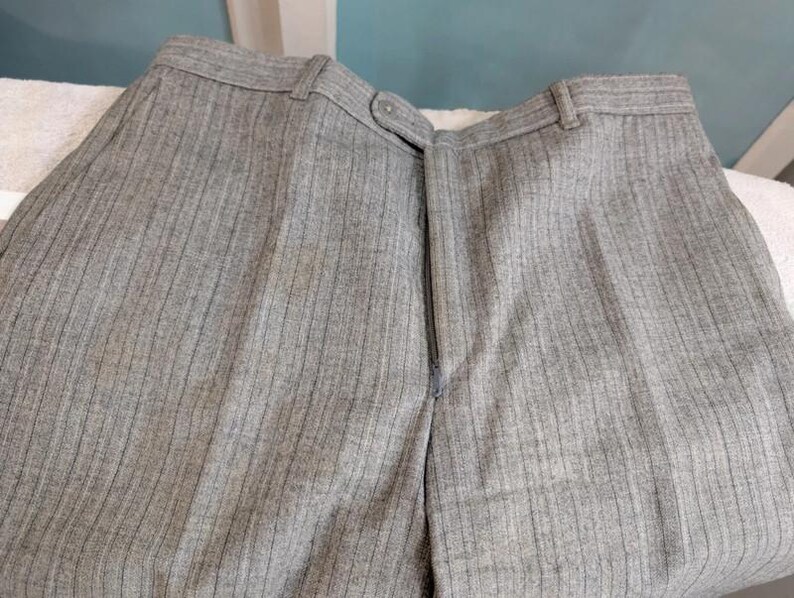 May include: Close-up of gray pinstripe trousers. The pants have a zipper closure and a button at the waistband. The fabric has vertical stripes. The trousers are made of a medium-weight fabric.