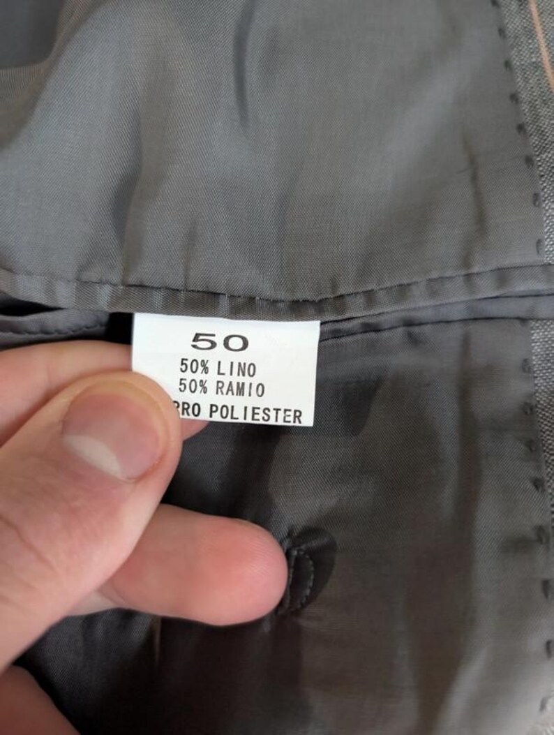 Może przedstawiać: Zbliżenie szarej podszewki ubrania z białą metką. Metka zawiera numer "50" oraz skład materiału: 50% LINO, 50% RAMIO i PRO POLIESTER. Ubranie wygląda na kurtkę lub marynarkę.