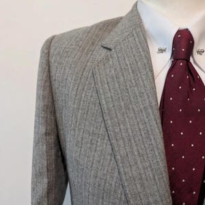 May include: A gray pinstripe suit jacket with a white dress shirt and a burgundy tie with white polka dots. The suit is displayed on a mannequin. The tie has a tie pin.