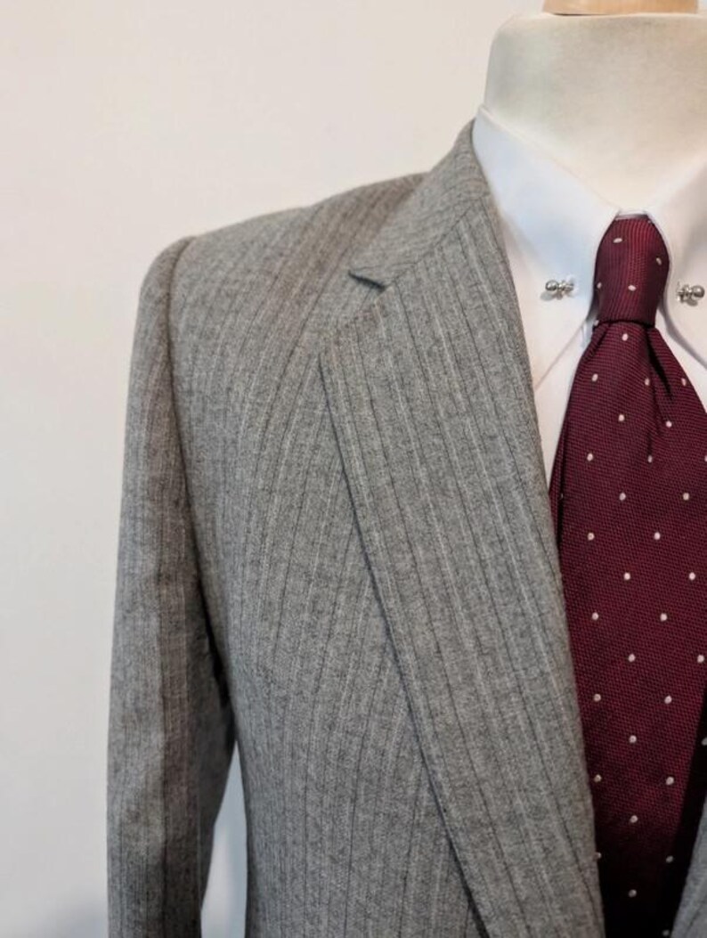 May include: A gray pinstripe suit jacket with a white dress shirt and a burgundy tie with white polka dots. The suit is displayed on a mannequin. The tie has a tie pin.