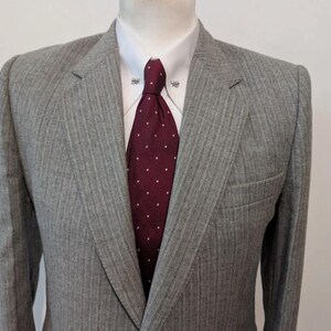 May include: A gray pinstripe suit jacket with a white dress shirt and a burgundy tie with white polka dots. The jacket has a notched lapel and a chest pocket. The tie is secured with a tie clip.