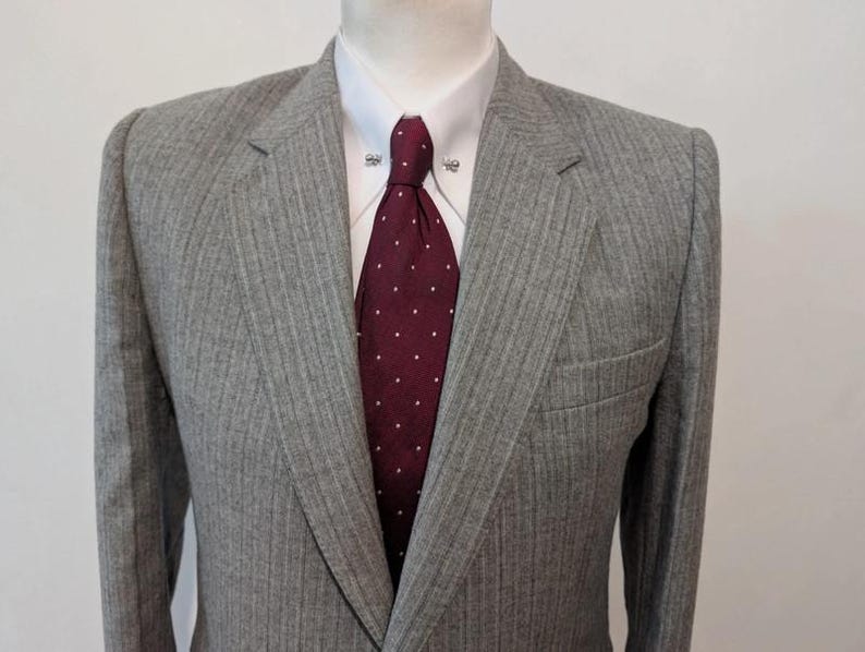 May include: A gray pinstripe suit jacket with a white dress shirt and a burgundy tie with white polka dots. The jacket has a notched lapel and a chest pocket. The tie is secured with a tie clip.