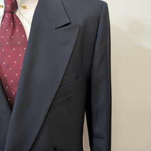 May include: A navy blue double-breasted suit jacket with a notched lapel. The jacket has a two-button closure and a welt pocket. A white dress shirt and a burgundy tie with white polka dots are visible.