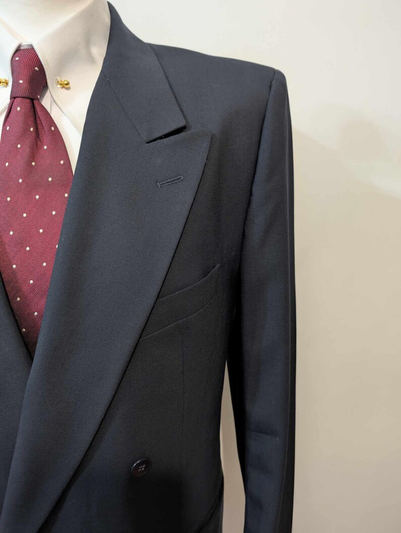 May include: A navy blue double-breasted suit jacket with a notched lapel. The jacket has a two-button closure and a welt pocket. A white dress shirt and a burgundy tie with white polka dots are visible.