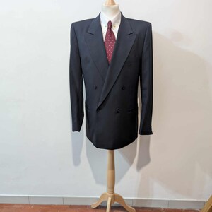 May include: A dark navy double-breasted blazer, white collared shirt, and burgundy patterned tie on a wooden mannequin. The blazer features a classic design with notched lapels and a double-button closure. The suit is a formal wear item.